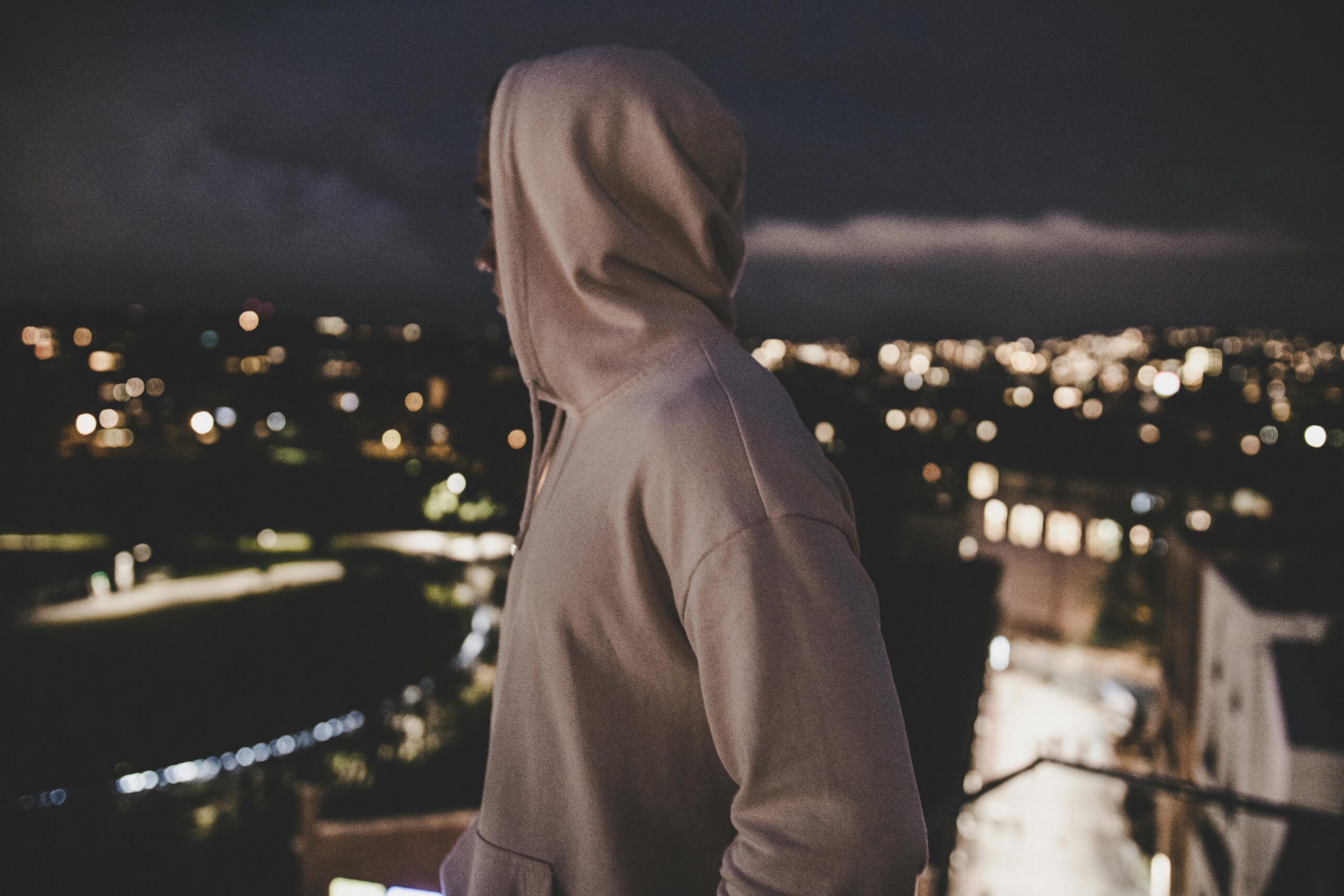 A person in a hoodie overlooks a blurry cityscape at night, evoking mystery and solitude.